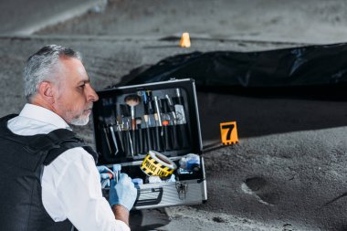ceset torbası içinde ceset ile olay yerinde inceleme araçlarıyla durumda oturan orta yaşlı polis