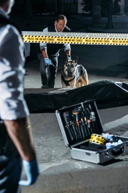 partial view of police officer standing near case with investigation tools while his colleague standing with german shepherd on leash near corpse at crime scene  clipart