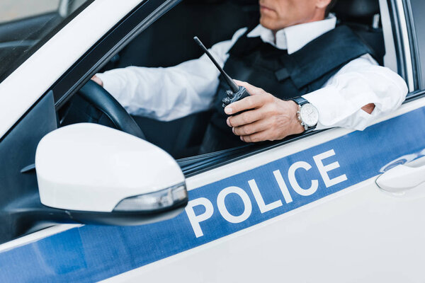 partial view of male police officer in bulletproof vest holding walkie-talkie in car 