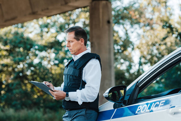 side view of middle aged male police officer in bulletproof vest writing in clipboard near car at street 