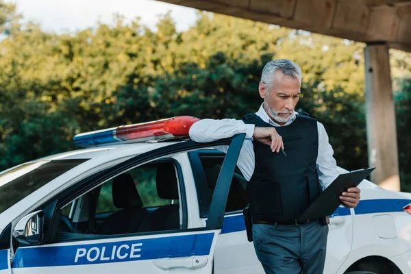 Volwassen Politieman Kogelvrij Vest Kijken Naar Klembord Buurt Van Auto — Stockfoto