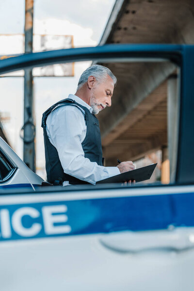 low angle view of mature policeman in bulletproof vest writing in clipboard near car at street