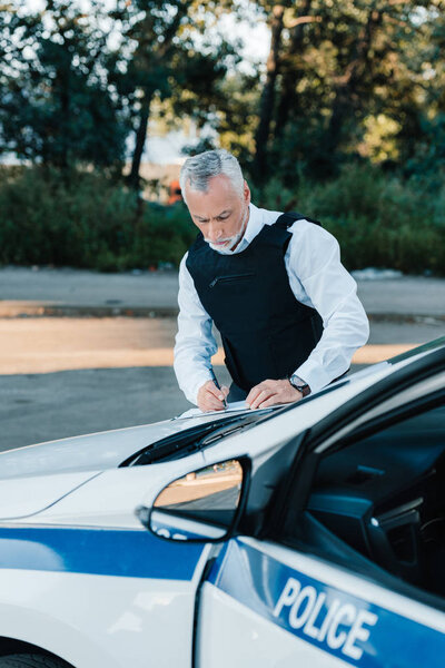 concentrated mature male police officer writing in clipboard on car at street