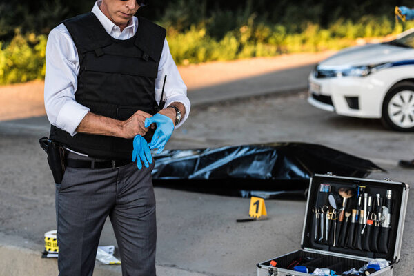 partial view of policeman with gun in holster putting on latex gloves at crime scene with corpse in body bag and case with investigation tools 