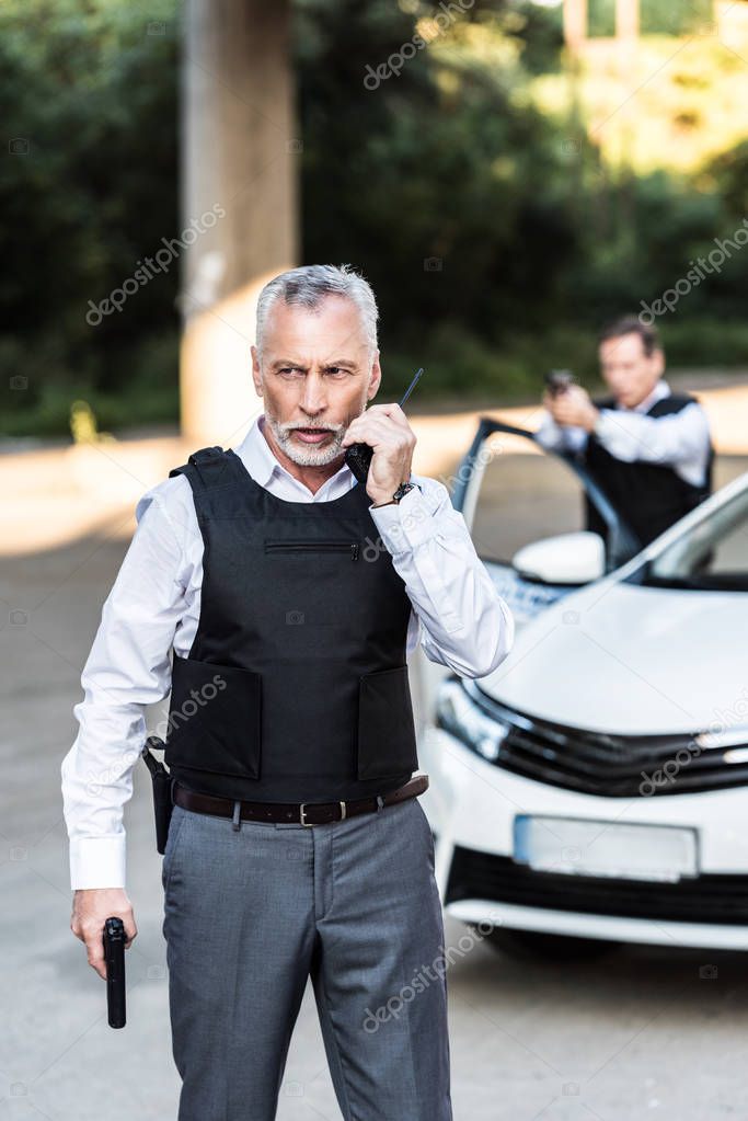 mature policeman with handgun talking on radio set while his colleague aiming by gun behind at street 