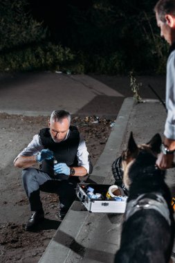 male police officer in latex gloves collecting evidence with case for investigation tools while his colleague standing with dog on leash at crime scene clipart