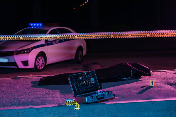 toned picture of crime scene with police car, case with investigation tools, cross line and corpse in body bag
