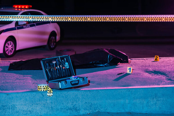 toned picture of case with investigation tools, cross line, police car and corpse in body bag at crime scene 