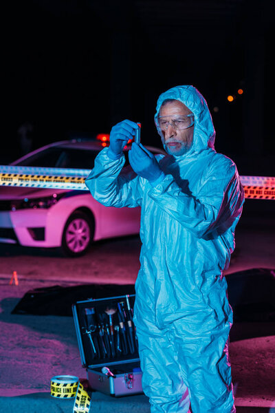 focused male criminologist in protective suit and latex gloves collecting evidence near cross line at crime scene 