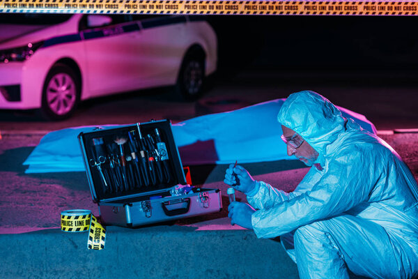 side view of male criminologist in protective suit and latex gloves putting evidence into flask by tweezers at crime scene with corpse 