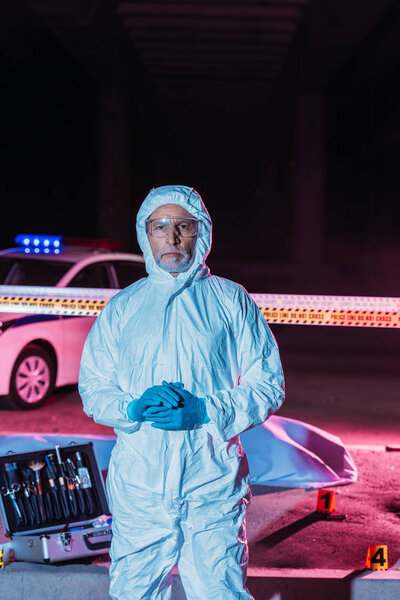 male criminologist in protective suit and mask looking at camera near crime scene with corpse in body bag and case with investigation tools 