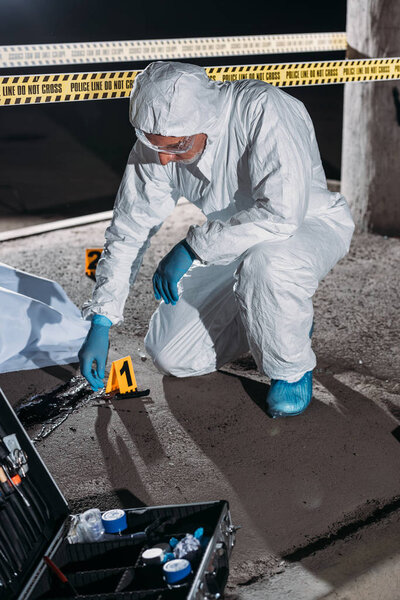 male criminologist in protective suit and latex gloves collecting evidence at crime scene with corpse 