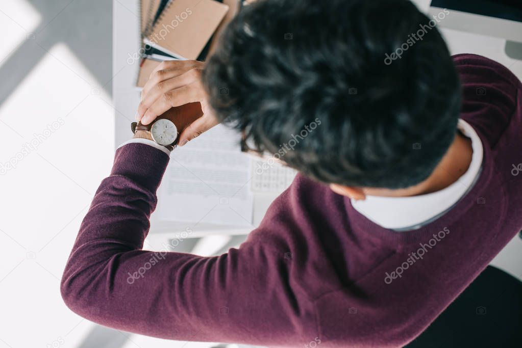 overhead view of businessman in burgundy sweater checking time at wristwatch in office