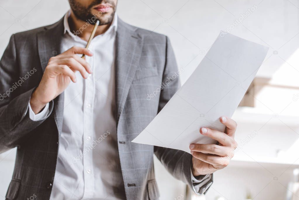 cropped image of businessman reading documents and touching chin with pencil in office