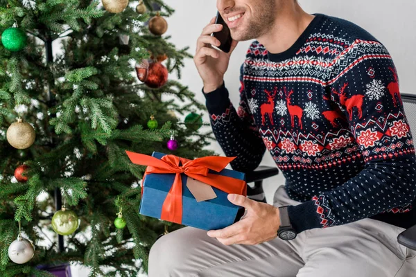 smiling man with gift sitting near christmas tree and talking on smartphone