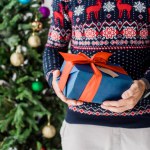 Vista recortada del hombre en suéter de Navidad con presente contra el árbol de Navidad