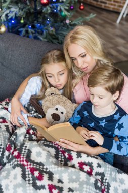 high angle view of young mother reading book with cute children in pajamas at christmas time 