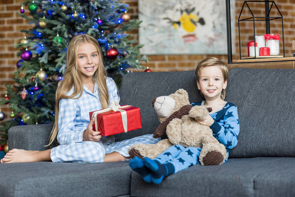 cute happy kids in pajamas holding teddy bear and christmas present while sitting in sofa and smiling at camera 
