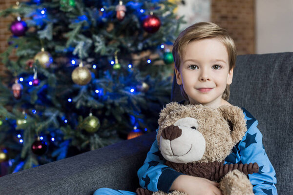 очаровательный счастливый ребенок обнимает плюшевого мишку и улыбается в камеру, сидя на диване на Рождество
