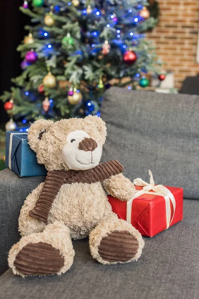 close-up view of teddy bear with gift boxes on sofa and decorated christmas tree behind