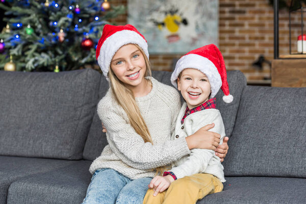 cute happy children in santa hats sitting together on couch and smiling at camera 