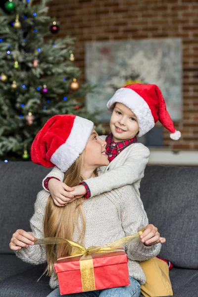 幸せな兄と妹サンタ帽子を抱き締めると クリスマス プレゼントを開くの — ストック写真