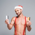 Young sexy muscular man in christmas hat showing credit cards isolated on grey background