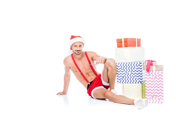 白い背景で隔離のギフト ボックスの山のそばに座ってクリスマス帽子で筋肉の上半身裸の男  — 無料ストックフォト