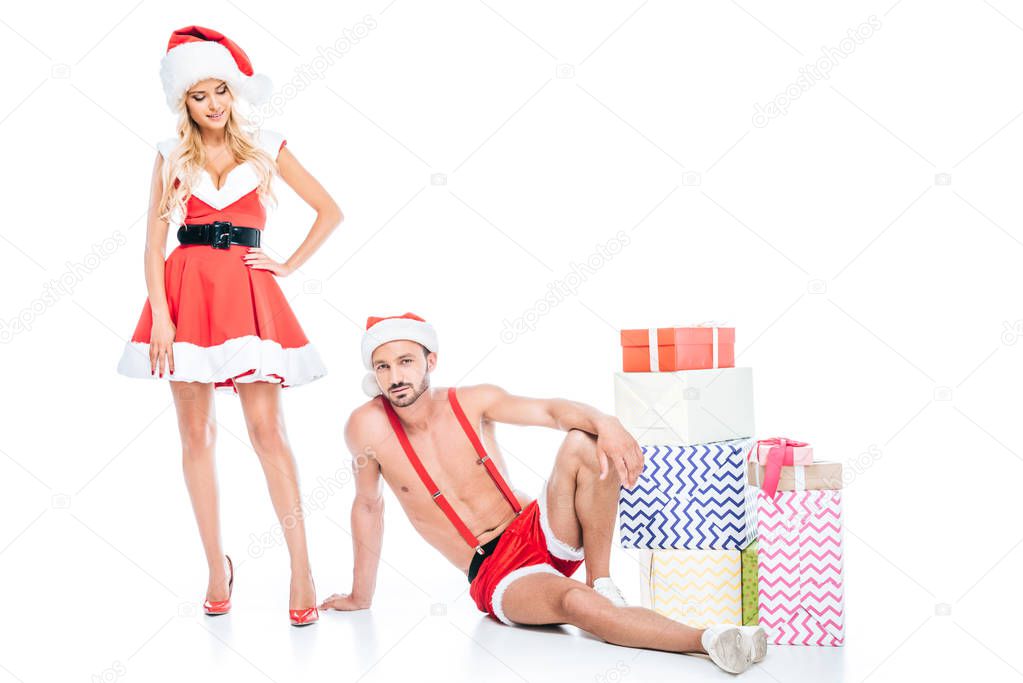 young muscular shirtless man in christmas hat laying on floor while his girlfriend standing near pile of gift boxes isolated on white background