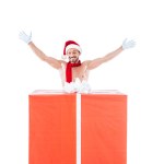 Happy shirtless muscular man in christmas hat standing with raised arms near big gift box isolated on white background