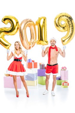 mutlu Noel Baba elbiseli Genç kadın ve kas gömleksiz sevgilisi Noel şapka işareti 2019 altın hava yapılan holding içinde hediye kutuları yığını önünde balonlar beyaz arka plan üzerinde izole 