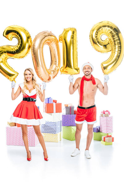 happy young woman in santa dress and her muscular shirtless boyfriend in christmas hat holding sign 2019 made of golden air balloons in front of pile of gift boxes isolated on white background 