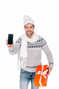 handsome young man in sweater, scarf and hat holding gift box and smartphone with blank screen isolated on white clipart