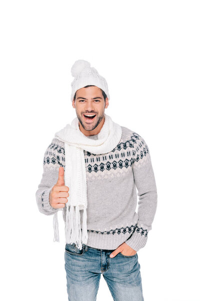 handsome young man in sweater, scarf and hat showing thumb up and smiling at camera isolated on white
