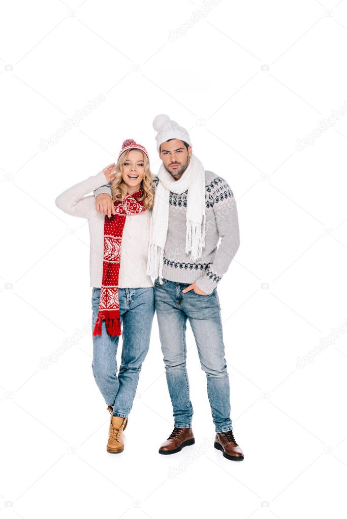 beautiful happy young couple in sweaters, scarves and hats standing together and looking at camera isolated on white
