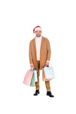 Alışveriş torbaları tutarak ve üzerinde beyaz izole kamera gülümseyen santa şapka yakışıklı sakallı genç adam
