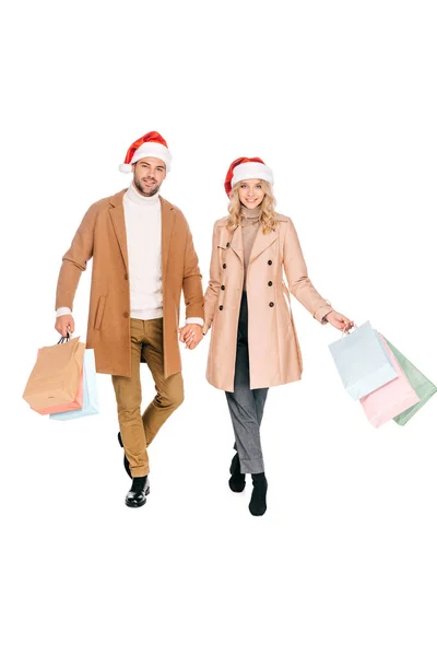 快乐的年轻夫妇在圣诞老人的帽子举行购物袋 微笑着在白色的相机隔离 — 图库照片