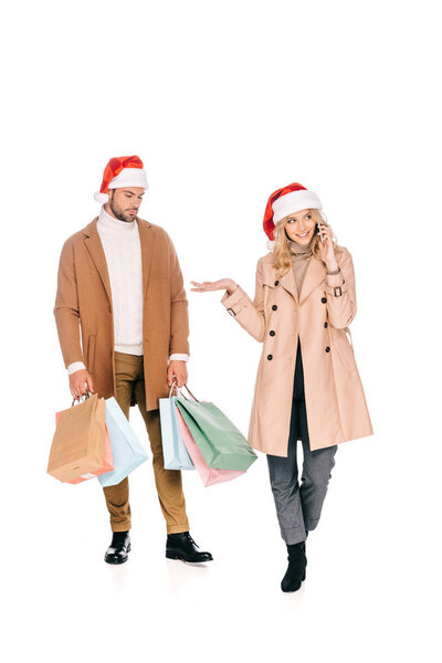 young man in santa hat holding shopping bags and looking at woman talking by smartphone isolated on white