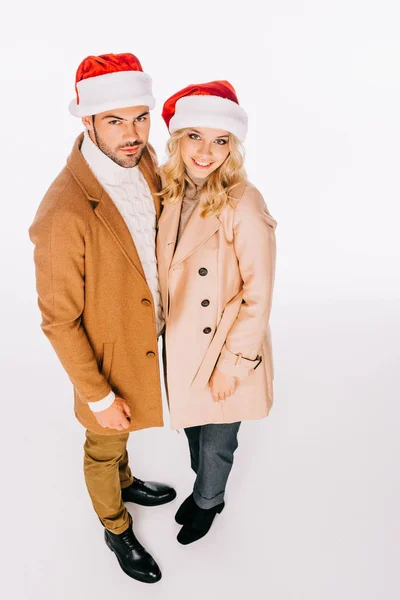 美丽的年轻夫妇的高角度视图在圣诞老人的帽子微笑着在白色的相机隔离 — 免费的图库照片