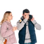 Sonriente joven con mochila sosteniendo una taza de papel y mirando a un hombre guapo fotografiando con cámara aislada en blanco