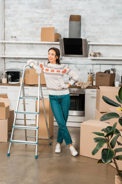 young woman in working gloves standing near ladder in kitchen with cardboard boxes at new home 