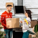 Knappe jonge man met kartonnen doos met kerstballen voor het verfraaien van de kerstboom terwijl zijn vriendin in de buurt van permanent in keuken thuis