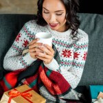 Vista ad alto angolo di giovane donna in possesso di tazza di caffè e seduta sul divano sotto coperta con scatole regalo di Natale a casa