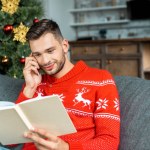 Joven guapo leyendo libro y hablando en el teléfono inteligente cerca del árbol de Navidad en casa