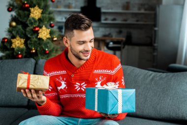kanepede oturan ve hediye kutuları Noel ağacı yakınındaki evde tutan gülümseyen genç adam 