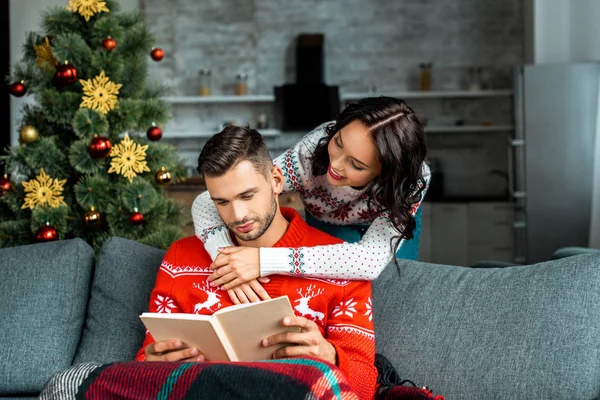 beautiful young woman hugging boyfriend from behind while he reading book on sofa near christmas tree at home