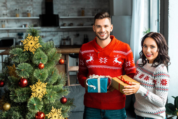 couple with gift boxes looking at camera near christmas tree at home 