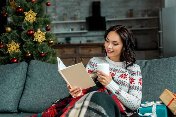 Smiling woman with coffee cup reading book on sofa under blanket with gifts near christmas tree at home — Stock Image Smiling Woman Coffee Cup Reading Book Sofa Blanket Gifts Christmas Stock Picture