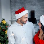 Sonriente pareja joven en sombreros de Navidad sosteniendo luces de bengala y mirándose en casa