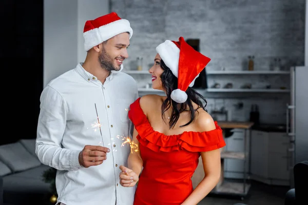 輝きと家庭でお互いを見てクリスマス帽子で幸せな若いカップル  — 無料ストックフォト